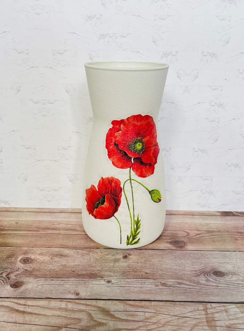 May include: White ceramic vase with a floral design featuring two red poppies. The vase has a textured surface and a slightly flared opening.
