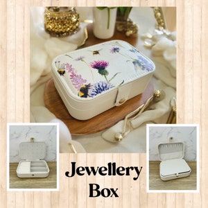 May include: A white jewellery box with a floral and bee design. The box has a hinged lid and a small compartment inside. The box is sitting on a wooden surface.