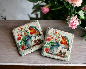 May include: Two square ceramic coasters with a robin bird design. Each coaster features a robin perched on a bucket with flowers and greenery. The background is a white watercolor wash with splashes of color.