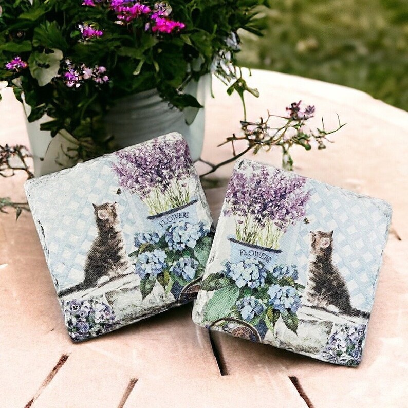 May include: Two square decorative coasters with a cat and floral design. Each coaster features a tabby cat, lavender, and blue hydrangeas in a blue flower pot. The background has a light blue checkered pattern.