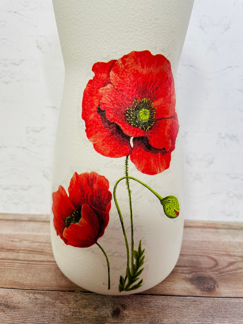 May include: White ceramic vase with a floral design featuring red poppies. The vase has a textured surface and a narrow neck.