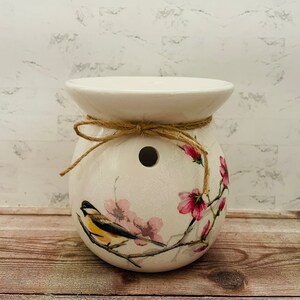 May include: White ceramic wax melt warmer with a floral design featuring a bird and pink flowers. The warmer has a hole in the top for a tea light and is tied with twine.