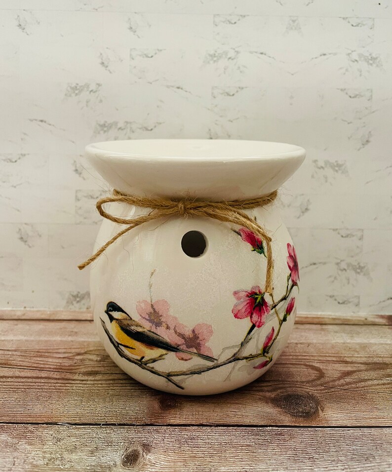 May include: White ceramic wax melt warmer with a floral design featuring a bird and pink flowers. The warmer has a hole in the top for a tea light and is tied with twine.