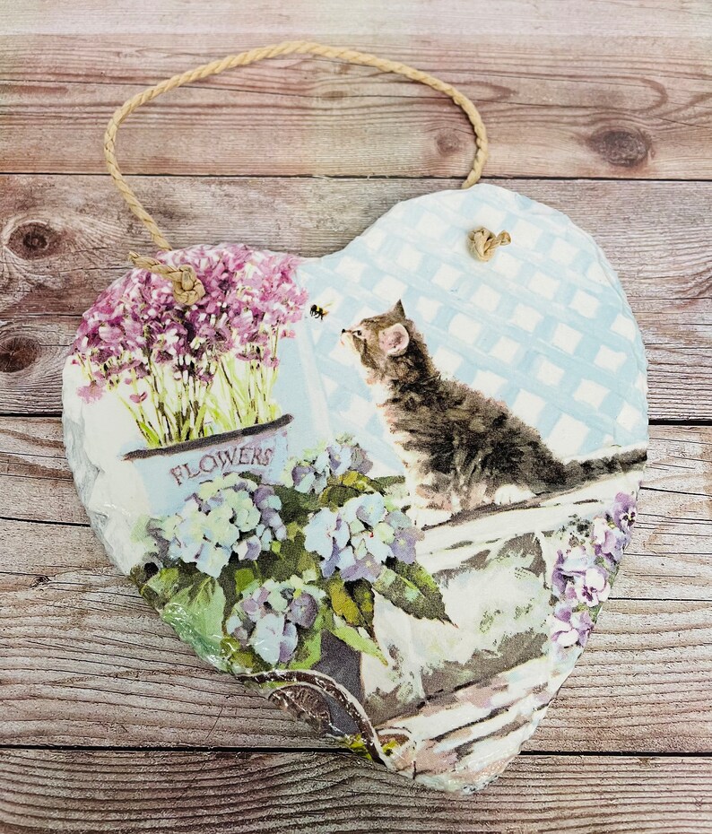 K&ouml;nnte beinhalten: Eine herzf&ouml;rmige Schiefertafel mit blau-wei&szlig;em kariertem Hintergrund. Die Tafel zeigt ein Blumenmuster mit einer Katze, die eine Biene betrachtet, und den Text "FLOWERS".