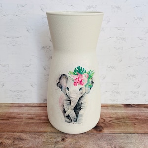 May include: White ceramic vase with a floral design featuring a gray elephant wearing a pink flower crown. The elephant is surrounded by green leaves and pink flowers.