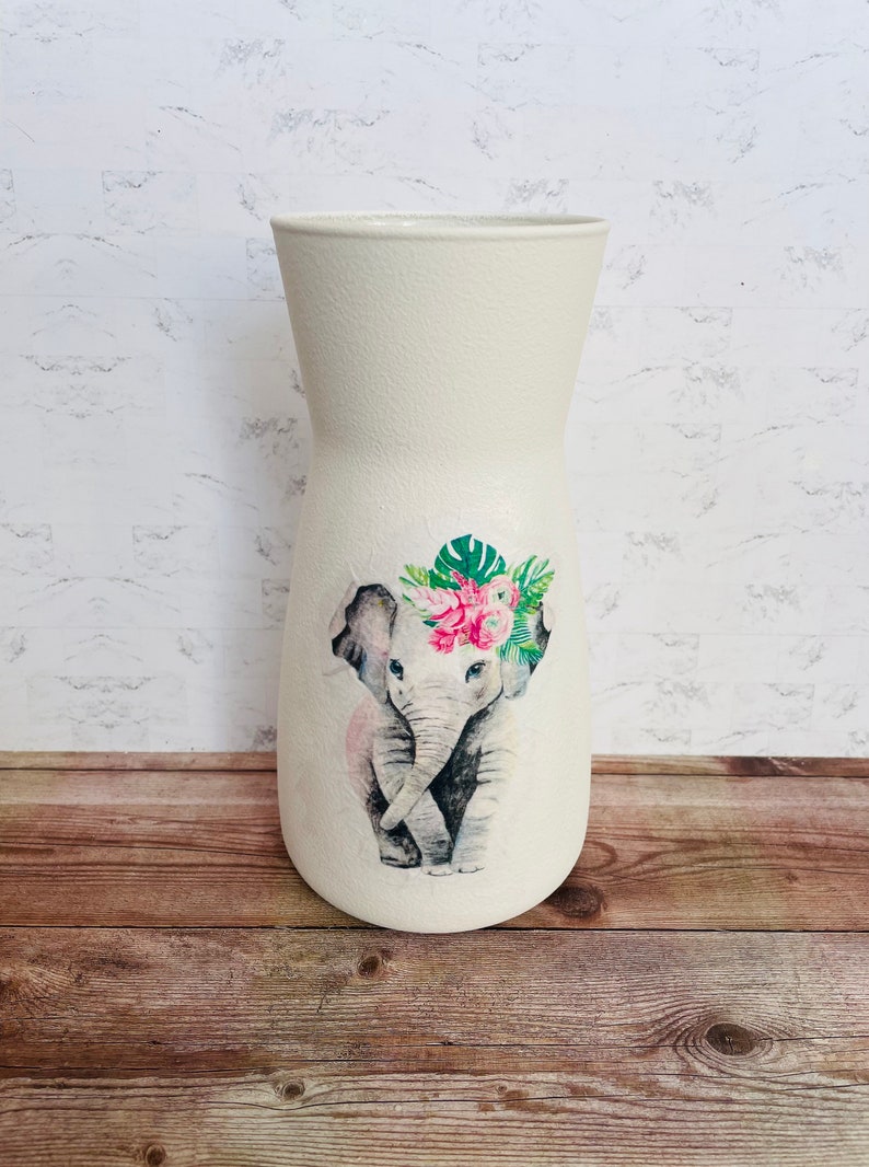 May include: White ceramic vase with a floral design featuring a gray elephant wearing a pink flower crown. The elephant is surrounded by green leaves and pink flowers.