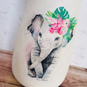 May include: A white ceramic vase with a watercolor illustration of an elephant wearing a floral crown. The elephant is gray with pink accents and the flowers are pink and green.
