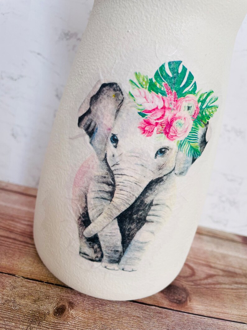 May include: A white ceramic vase with a watercolor illustration of an elephant wearing a floral crown. The elephant is gray with pink accents and the flowers are pink and green.