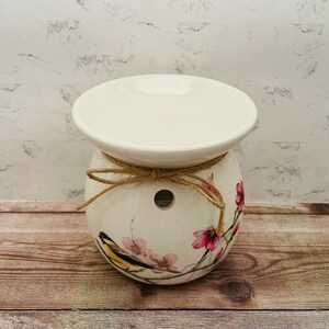 May include: White ceramic wax warmer with a floral design featuring pink flowers and a yellow bird. The warmer has a top plate for melting wax and a hole in the bottom for a tea light.