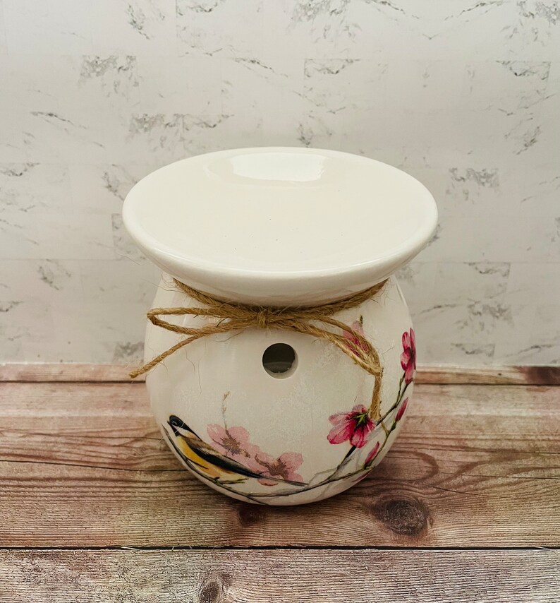 May include: White ceramic wax warmer with a floral design featuring pink flowers and a yellow bird. The warmer has a top plate for melting wax and a hole in the bottom for a tea light.