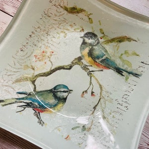 May include: A light blue glass serving tray with a curved edge features a watercolor design of two bluebirds perched on a branch with pink flowers. The background is a light gray with a faded script pattern.