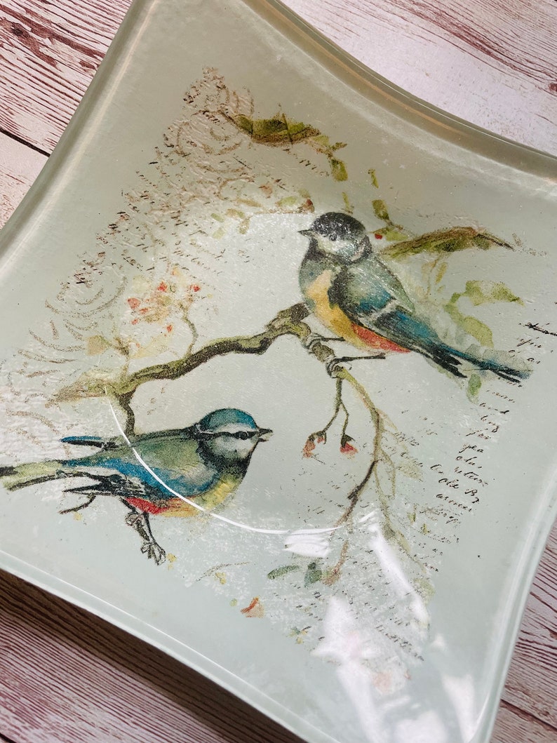 May include: A light blue glass serving tray with a curved edge features a watercolor design of two bluebirds perched on a branch with pink flowers. The background is a light gray with a faded script pattern.