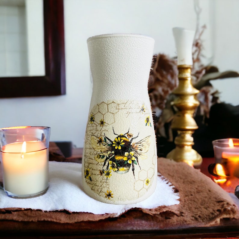 K&ouml;nnte beinhalten: Eine wei&szlig;e Keramikvase mit strukturierter Oberfl&auml;che zeigt ein Bienen- und Wabenmuster. Die Biene ist gelb und schwarz mit braunen Fl&uuml;geln und von gelben Blumen umgeben. Die Vase hat einen schmalen Hals und einen breiten Boden.