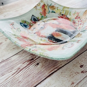 Pu&ograve; includere: Un piatto da portata quadrato in vetro con un design ad acquerello di una volpe in un ambiente floreale. Il piatto ha uno sfondo verde chiaro con accenti rosa, bianchi e marroni.
