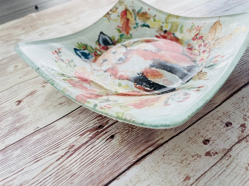 Pu&ograve; includere: Un piatto da portata quadrato in vetro con un design ad acquerello di una volpe in un ambiente floreale. Il piatto ha uno sfondo verde chiaro con accenti rosa, bianchi e marroni.