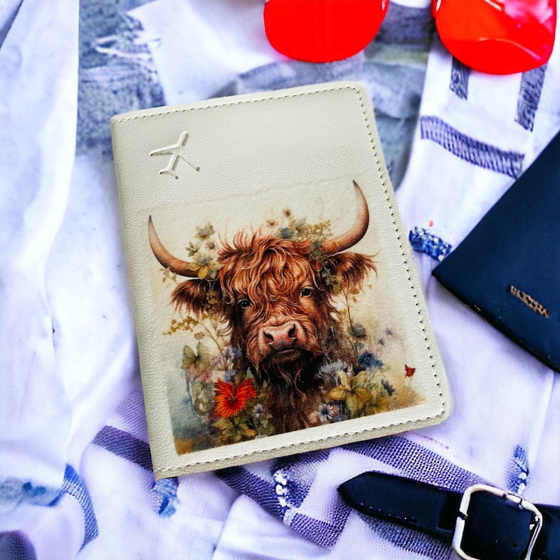 Pu&ograve; includere: Una custodia per passaporto beige con un'illustrazione di una mucca Highland marrone e bianca. La mucca &egrave; circondata da fiori e farfalle. La custodia ha una piccola icona di aereo nell'angolo in alto a sinistra.