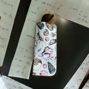 May include: A white sunglasses case featuring a seashell design. The case has a zip fastening and is made from a hard-wearing material.