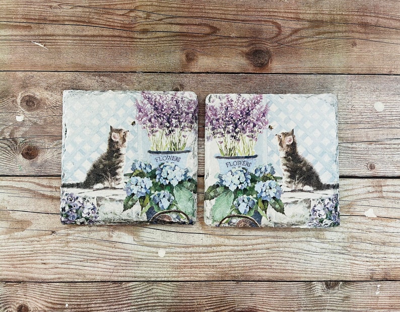 May include: Two square decorative tiles with a vintage design. Each tile features a kitten, lavender, and blue hydrangeas. The word "FLOWERS" is printed on the pots. The background has a light blue trellis pattern.