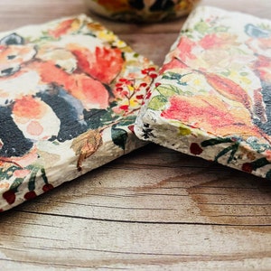 May include: Two square coasters with a watercolor design of a fox in a forest setting. The coasters feature a white background with red, orange, green, and brown accents. The fox is depicted in a standing position with its tail curled around its body.
