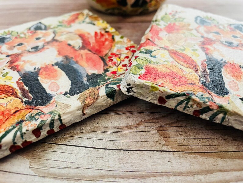 May include: Two square coasters with a watercolor design of a fox in a forest setting. The coasters feature a white background with red, orange, green, and brown accents. The fox is depicted in a standing position with its tail curled around its body.