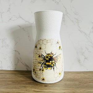 K&ouml;nnte beinhalten: Wei&szlig;e Keramikvase mit strukturierter Oberfl&auml;che, die eine Biene mit gelben und schwarzen Streifen und Blumen zeigt. Die Biene ist von einem Wabenmuster und einem Vintage-Blumendesign umgeben.