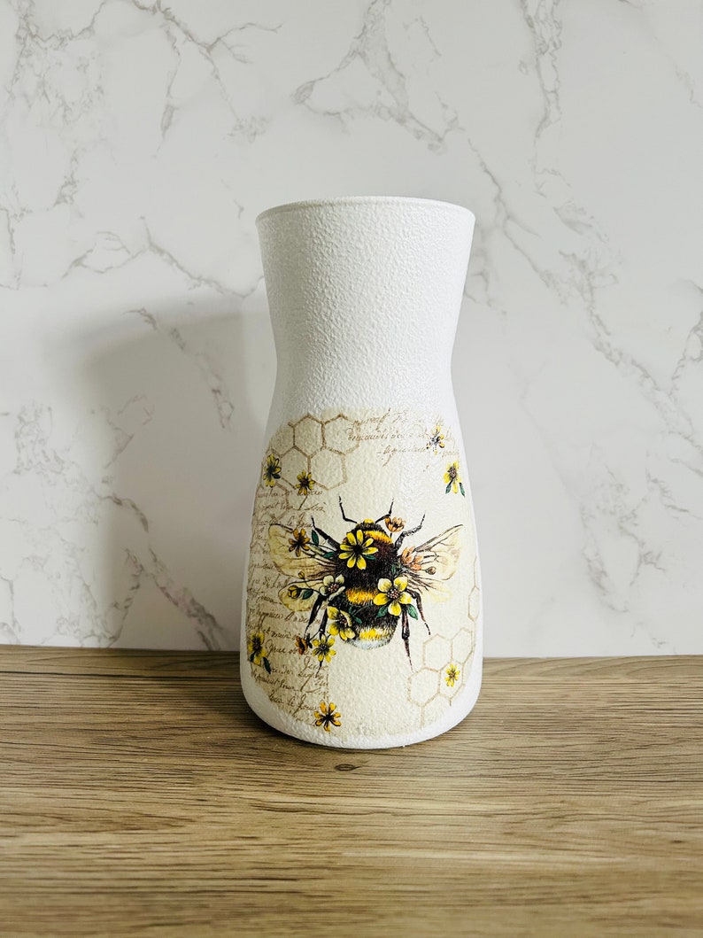 K&ouml;nnte beinhalten: Wei&szlig;e Keramikvase mit strukturierter Oberfl&auml;che, die eine Biene mit gelben und schwarzen Streifen und Blumen zeigt. Die Biene ist von einem Wabenmuster und einem Vintage-Blumendesign umgeben.