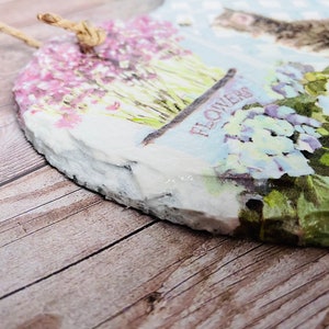 K&ouml;nnte beinhalten: Eine herzf&ouml;rmige Schiefertafel mit einem Blumenmuster. Die Tafel zeigt eine Vielzahl von Blumen, darunter rosa, lila und wei&szlig;e Bl&uuml;ten. Die Tafel ist mit einem rustikalen, abgenutzten Look verziert und hat einen Bindfaden zum Aufh&auml;ngen.