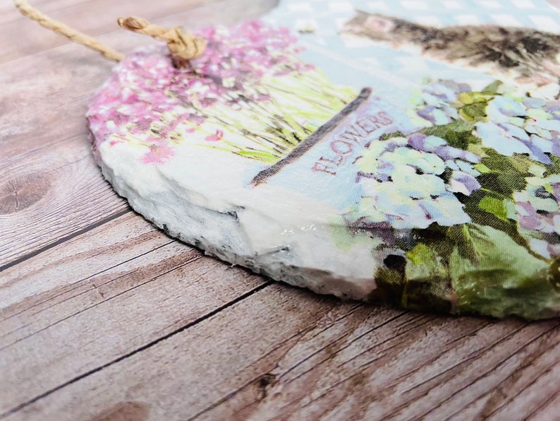 K&ouml;nnte beinhalten: Eine herzf&ouml;rmige Schiefertafel mit einem Blumenmuster. Die Tafel zeigt eine Vielzahl von Blumen, darunter rosa, lila und wei&szlig;e Bl&uuml;ten. Die Tafel ist mit einem rustikalen, abgenutzten Look verziert und hat einen Bindfaden zum Aufh&auml;ngen.