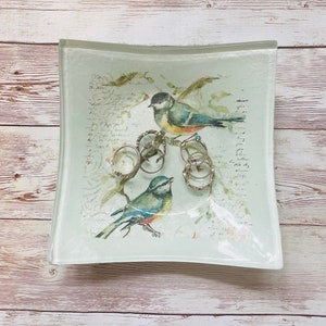 May include: A white glass trinket dish with a floral design and two blue birds. The dish is filled with silver rings.