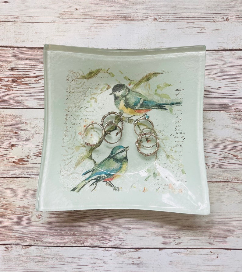 May include: A white glass trinket dish with a floral design and two blue birds. The dish is filled with silver rings.