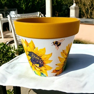 May include: A yellow terracotta flower pot with a white background featuring a sunflower and bee design.