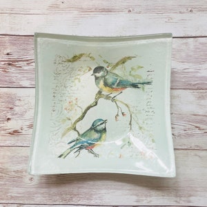 May include: A light blue glass dish with a curved edge features a watercolor design of two blue birds perched on a branch with pink flowers. The background is a light blue with a faded white script.