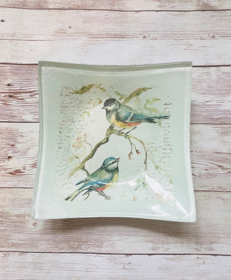 May include: A light blue glass dish with a curved edge features a watercolor design of two blue birds perched on a branch with pink flowers. The background is a light blue with a faded white script.
