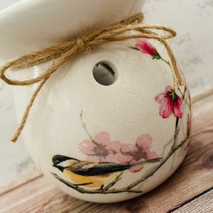 May include: White ceramic wax melt warmer with a floral design featuring pink flowers and a small bird. The warmer has a hole in the top for a wax melt and is tied with a brown twine bow.