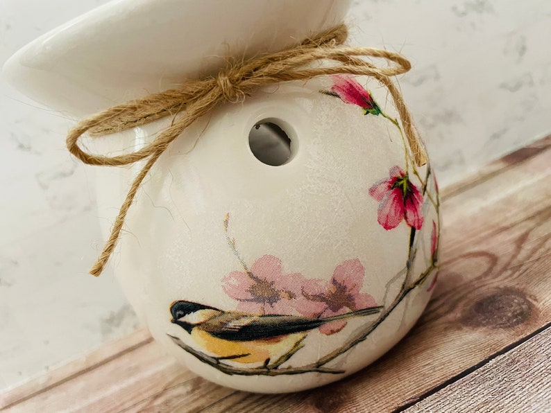 May include: White ceramic wax melt warmer with a floral design featuring pink flowers and a small bird. The warmer has a hole in the top for a wax melt and is tied with a brown twine bow.