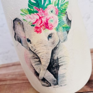 May include: A watercolor illustration of a baby elephant wearing a floral crown with pink and green flowers. The elephant is gray and has a pink nose. The background is white.