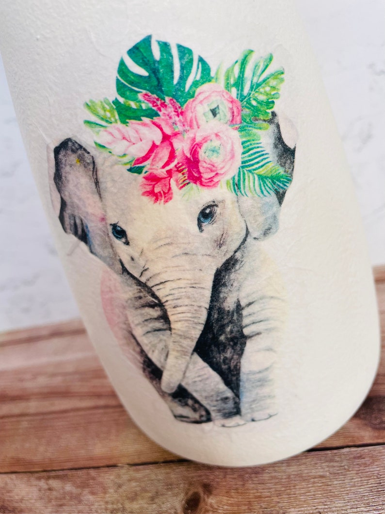 May include: A watercolor illustration of a baby elephant wearing a floral crown with pink and green flowers. The elephant is gray and has a pink nose. The background is white.