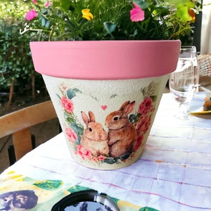 May include: A pink terracotta planter with a white background featuring a floral design and two brown rabbits. The rabbits are facing each other and have a small heart between them.