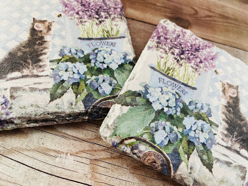 May include: Two decorative stone tiles with a floral and cat design. The tiles feature a watercolor-style print of a kitten, blue hydrangeas, and lavender in a white flower pot. The word "FLOWERS" is printed on the flower pot.