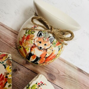 May include: A white ceramic wax melt warmer with a fox and floral design. The warmer has a brown twine bow and is sitting on a wooden surface.