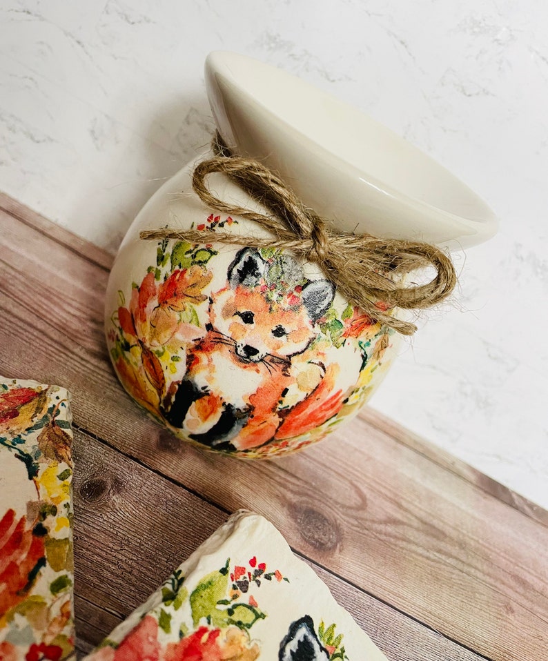 May include: A white ceramic wax melt warmer with a fox and floral design. The warmer has a brown twine bow and is sitting on a wooden surface.