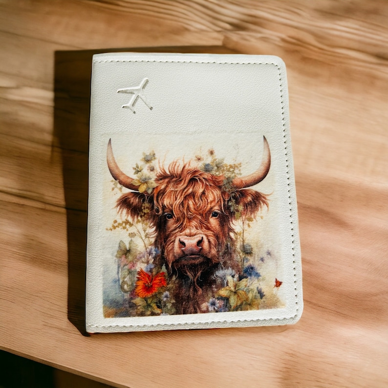 Pu&ograve; includere: Porta passaporto bianco con un'illustrazione di una mucca delle Highlands marrone e bianca circondata da fiori. Il porta passaporto ha una piccola icona di aereo sul davanti.