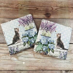 May include: Two square decorative coasters with a cat and floral design. Each coaster features a tabby cat, blue hydrangeas, and lavender in a blue flower pot. The background has a light blue checkered pattern.