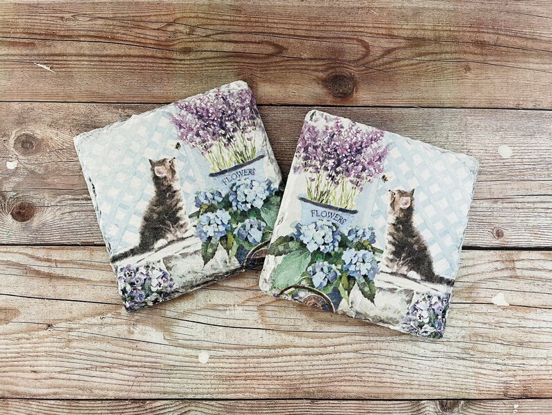 May include: Two square decorative coasters with a cat and floral design. Each coaster features a tabby cat, blue hydrangeas, and lavender in a blue flower pot. The background has a light blue checkered pattern.