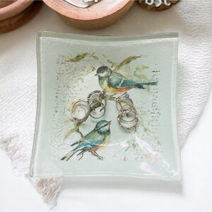 May include: A clear glass trinket dish with a square shape and rounded edges. The dish features a printed design of two birds perched on a branch with a floral background. The design includes handwritten text and several rings are placed on the branch.