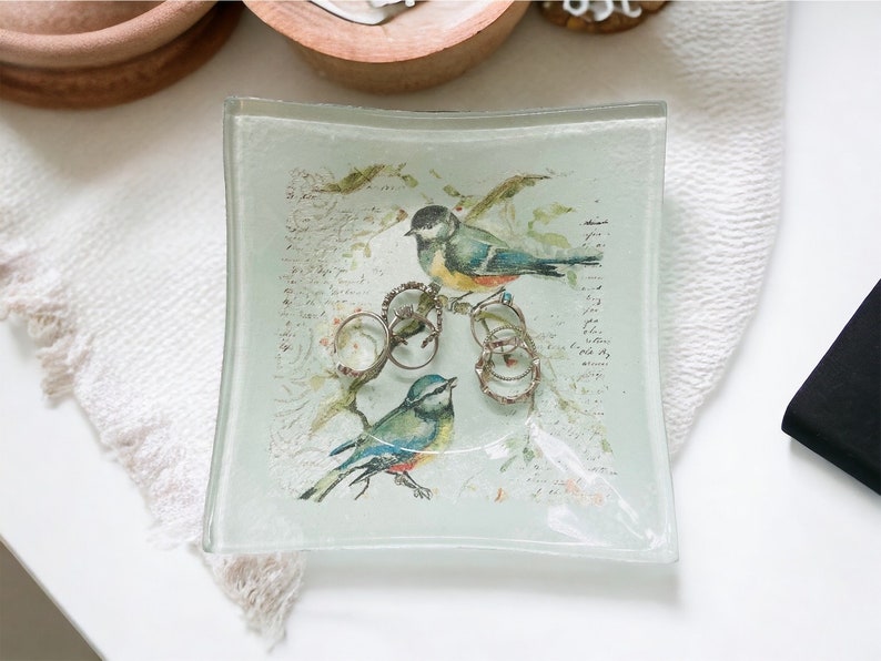 May include: A clear glass trinket dish with a square shape and rounded edges. The dish features a printed design of two birds perched on a branch with a floral background. The design includes handwritten text and several rings are placed on the branch.