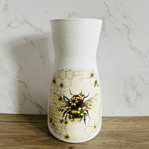 K&ouml;nnte beinhalten: Wei&szlig;e Keramikvase mit strukturierter Oberfl&auml;che. Die Vase zeigt eine Biene mit gelben und schwarzen Streifen und Blumen auf einem Honigwabenhintergrund.