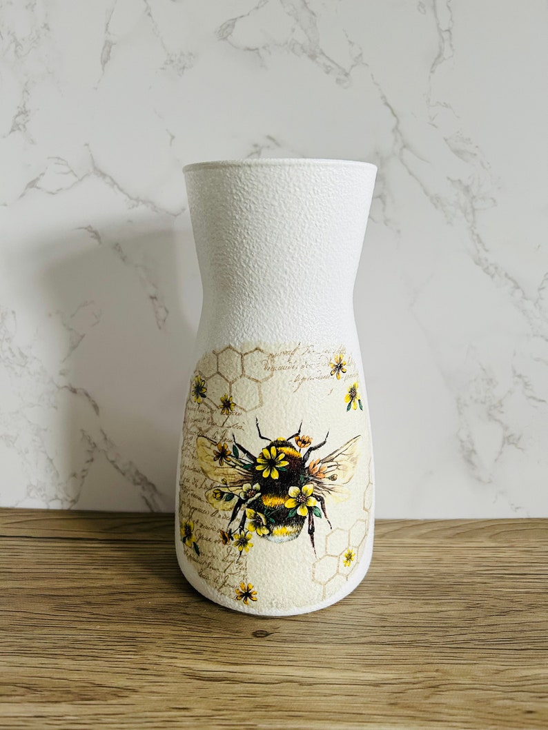 K&ouml;nnte beinhalten: Wei&szlig;e Keramikvase mit strukturierter Oberfl&auml;che. Die Vase zeigt eine Biene mit gelben und schwarzen Streifen und Blumen auf einem Honigwabenhintergrund.