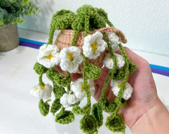 May include: A crocheted hanging plant with white flowers and green leaves. The plant is in a tan pot and has long, trailing vines with white flowers and yellow centers. The leaves are green and the pot is round.