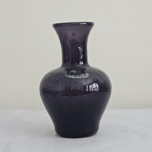 May include: A dark purple glass vase with a rounded body and a flared neck. The vase has a textured surface and reflects light, creating highlights and shadows. The vase is a decorative item.